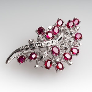 May include: A silver-toned brooch featuring a floral design with numerous oval-cut, deep red gemstones. The brooch has a curved, flowing shape, with smaller clear stones interspersed throughout the design.