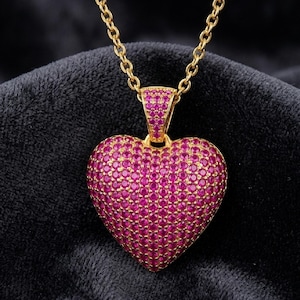 May include: A gold-toned necklace featuring a heart-shaped pendant completely covered in small, vibrant pink stones. The pendant is attached to a matching gold-toned chain, and the entire surface of the heart and the bail is encrusted with stones.