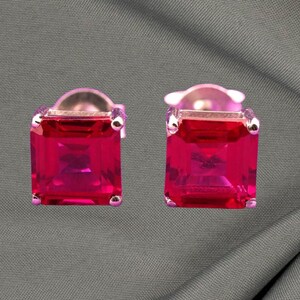 May include: A pair of square-cut, bright pink gemstone stud earrings. The gems are set in a silver-colored metal with small, round accents at each corner. The earrings are displayed against a gray background.