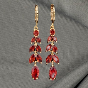 May include: Gold-coloured drop earrings with a leaf-inspired design. Each earring features a series of red, marquise-cut gemstones, with a larger, teardrop-shaped stone at the base. The earrings are suspended from a gold-coloured clasp.