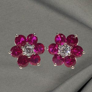 May include: A pair of flower-shaped stud earrings. Each earring features six magenta-colored gemstones surrounding a central clear gemstone. The earrings are set in a silver-colored metal.