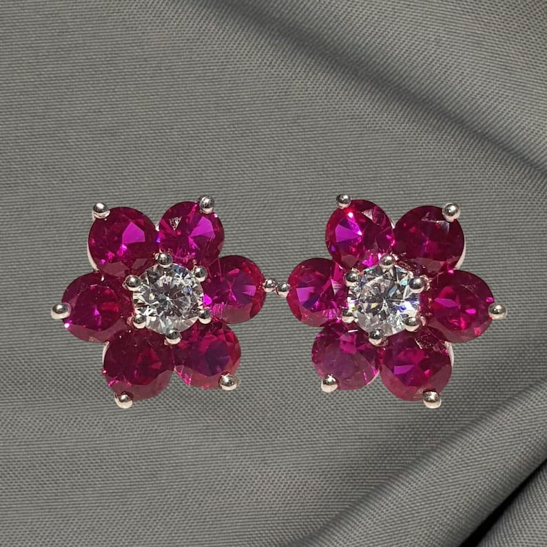 May include: A pair of flower-shaped stud earrings. Each earring features six magenta-colored gemstones surrounding a central clear gemstone. The earrings are set in a silver-colored metal.