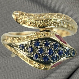 May include: A gold-toned ring in a snake design, featuring a central section of blue gemstones and accents of smaller clear stones. The ring has a curved, layered design with ruby-colored accents.