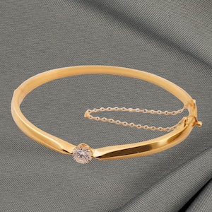 May include: A gold bangle bracelet with a single, clear gemstone. The bracelet has a simple, elegant design with a small safety chain. The background is a grey fabric.