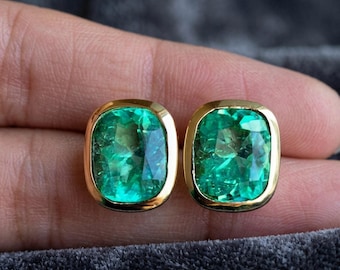 May include: A pair of stud earrings featuring cushion-cut, vibrant green gemstones set in polished gold bezels. The stones exhibit a faceted surface that reflects light, showcasing a deep emerald hue within the smooth, rounded gold frames.