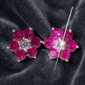May include: A pair of flower-shaped stud earrings featuring six vibrant fuchsia-pink round-cut stones arranged as petals around a central clear white round-cut stone, all set in a silver-toned metal frame with small prongs securing each gemstone.