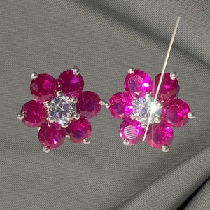 May include: A pair of flower-shaped earrings. Each earring features five vibrant pink gemstones arranged around a central, sparkling white gemstone. The earrings have a silver-colored metal setting.