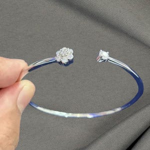 May include: A silver-coloured open cuff bracelet. One end features a cluster of small, round, clear stones in a floral arrangement. The other end has a single, larger, clear, round stone. The bracelet is held by a hand against a grey background.