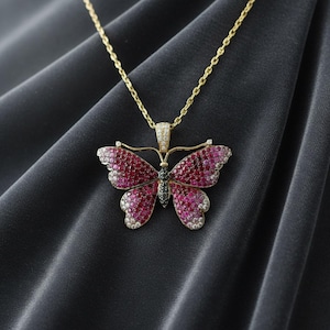 May include: A gold necklace featuring a butterfly pendant encrusted with numerous small red, pink, and white gemstones. The butterfly body is accented with dark stones, and the pendant hangs from a delicate gold chain link necklace.