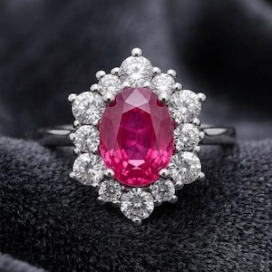 May include: A silver-toned ring featuring a large, oval-cut vibrant red gemstone at the center. The central stone is surrounded by a halo of twelve round, clear, faceted stones arranged in a floral pattern, set on a polished metal band.