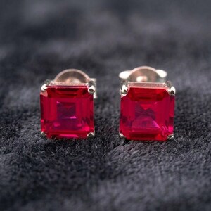 May include: A pair of stud earrings featuring square-cut, vibrant red gemstones set in polished silver-toned metal. Each earring displays a faceted surface that reflects light, secured with a standard post backing for attachment to the earlobe.