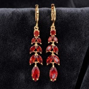May include: A pair of gold-toned drop earrings featuring a vertical arrangement of marquise-cut red gemstones. Each earring consists of a small round stone at the top, followed by three pairs of marquise stones, and a single larger marquise stone at the base.