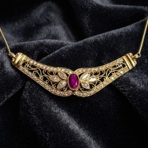 May include: A gold necklace featuring a curved, filigree-style centrepiece adorned with a central oval-cut magenta gemstone and numerous small, clear, round-cut stones set along the intricate metalwork, attached to a thin gold chain.
