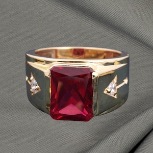 May include: A gold ring featuring a large, rectangular, ruby-red gemstone. The ring has a dark grey band with arrow and diamond accents. The ring is set against a grey background.