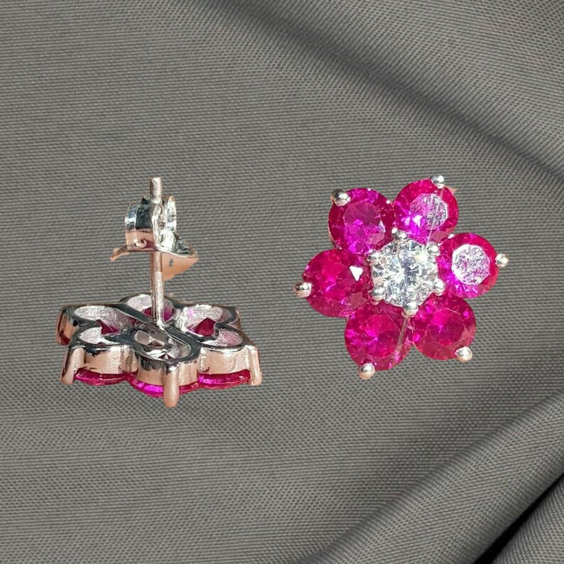 May include: A pair of flower-shaped stud earrings. Each earring features six vibrant pink gemstones surrounding a central, clear gemstone. The earrings are set in a silver-colored metal, creating a sparkling floral design.