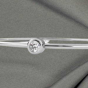 May include: A silver bangle bracelet with a single, round, clear diamond set in a bezel. The bracelet is thin and polished, with a simple, elegant design. The diamond is the focal point, catching the light and sparkling against the gray background.