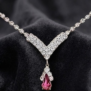 May include: A silver-toned necklace featuring a V-shaped centerpiece with a textured, hammered finish. A single, pear-shaped, deep pink gemstone pendant hangs from the center of the V-shape. The chain consists of small, reflective, faceted silver links.