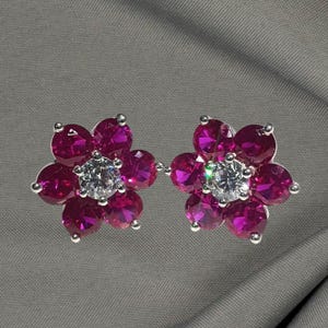May include: A pair of flower-shaped earrings. Each earring features five magenta-colored gemstones arranged around a central, clear, round gemstone. The earrings have silver-colored settings and are displayed on a gray background.