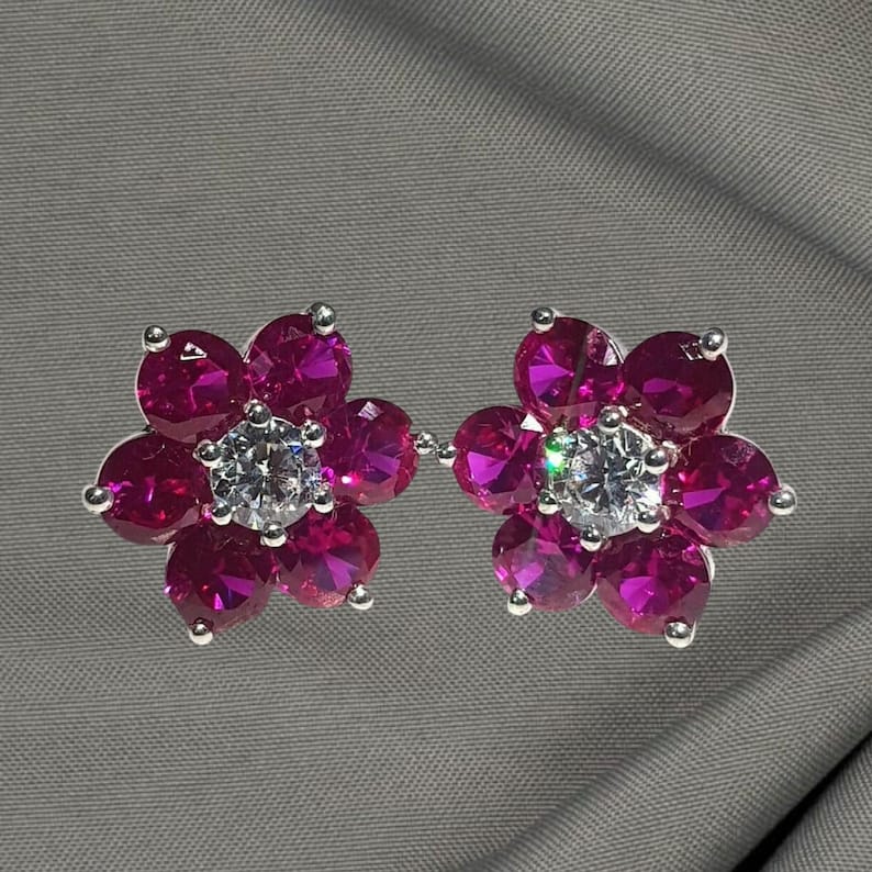 May include: A pair of flower-shaped earrings. Each earring features five magenta-colored gemstones arranged around a central, clear, round gemstone. The earrings have silver-colored settings and are displayed on a gray background.
