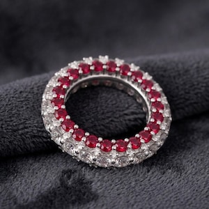 May include: A silver ring with a double row of gemstones. The inner row features small, round, red gemstones, while the outer row is composed of clear, white gemstones. The ring is set against a dark gray background.