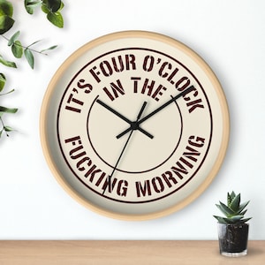 Può includere: Un orologio da parete rotondo con cornice in legno chiaro e quadrante color crema. Il centro presenta una scritta rosso scuro in stile usurato che recita IT'S FOUR O'CLOCK IN THE FUCKING MORNING disposta in cerchio con lancette nere.