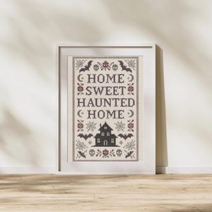 Può includere: Un'opera d'arte a punto croce incorniciata con le parole "HOME SWEET HAUNTED HOME" in viola scuro. Il design include pipistrelli, ragnatele, una casa infestata e accenti floreali, il tutto su uno sfondo color crema. La cornice è bianca.