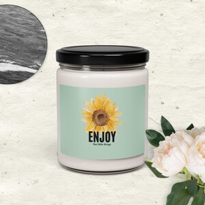 May include: A white candle jar with a black lid. The label features a yellow sunflower illustration on a light green background, with the word "ENJOY" and the phrase "the little things" in black text. A bouquet of white flowers is in the background.