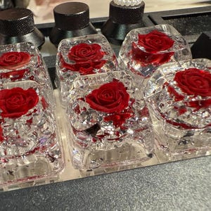 May include: Clear resin keyboard keycaps with embedded red roses. The keycaps are rectangular with rounded edges, and the roses are in full bloom. The keycaps are arranged on a keyboard, with other black and pink keys visible.
