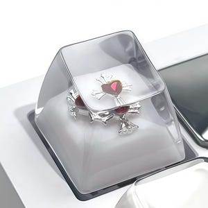 May include: A custom keyboard keycap featuring a silver cross with a red heart-shaped gem, encased in a clear, protective cover. The keycap is set on a white keyboard base, with other white and gray keycaps visible.