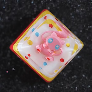 May include: A square-shaped resin art piece featuring a pink cartoon character with blue eyes. The piece has a white base with red, yellow, and blue accents. The background is a dark, glittery surface.