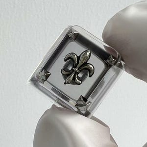 May include: A clear, square ring featuring a silver fleur-de-lis emblem. The ring has a silver border with small, pointed details. The ring is held by a gloved hand against a white backdrop.