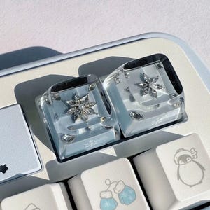 May include: Two clear, square keyboard keycaps with a snowflake design and embedded crystals. The keycaps are on a light blue keyboard, with other keys featuring winter-themed illustrations like mittens and a penguin.