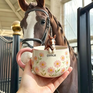 Może przedstawiać: Ceramiczny kubek z różowym uchem i kwiatowym wzorem różowych stokrotek na kremowym tle. Słowo COOKIE jest nadrukowane na czarno z przodu. Mała, trójwymiarowa figurka brązowo-białego konia jest przymocowana do wewnętrznej krawędzi naczynia.