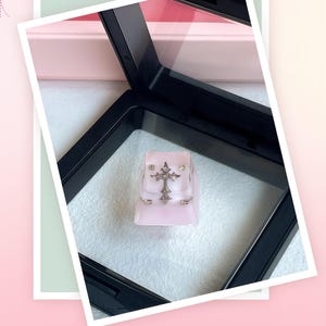 May include: A small, light pink backpack with silver-colored accents and a decorative cross on the front. The bag has a rectangular shape with a flap closure and small side pockets. The background is a soft white fabric.