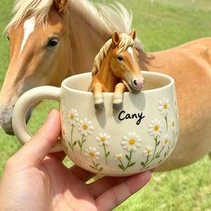 May include: A cream-coloured ceramic mug featuring a three-dimensional brown horse figurine emerging from the rim. The exterior is decorated with embossed white daisy flowers with yellow centres and green stems. The name Cany is printed in black.