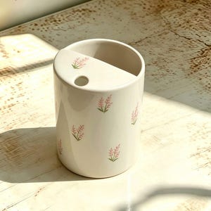 Könnte beinhalten: Ein zylindrischer Zahnbürstenhalter aus weißer Keramik mit einer kleinen kreisförmigen Öffnung auf der oberen Ablage und einem sich wiederholenden Muster aus kleinen rosa Blumenzweigen mit grünen Stielen auf der Außenfläche.