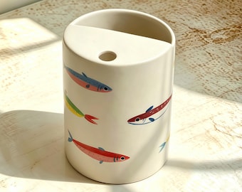 May include: A cylindrical off-white ceramic toothbrush holder featuring a pattern of small, colorful fish in shades of red, blue, green, and yellow. The top surface has a semi-circular opening and a single circular hole for holding items.