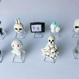 May include: A collection of Halloween-themed figurines, including a black tree, skull, television with handprints, ghosts, mummy, skeleton, and a gray rabbit with a red bow tie. Each figurine is displayed on a clear stand.