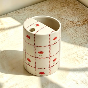 Könnte beinhalten: Ein zylindrischer Keramikbehälter mit cremeweißer Basis, verziert mit einem roten, wellenförmigen Gittermuster und kleinen roten Blumenmotiven. Die Oberseite ist teilweise mit einem flachen Abschnitt bedeckt, der ein einzelnes Loch aufweist.