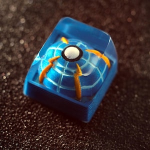 Peut inclure: Une touche artisanale bleue et orange avec un design sphérique. La touche présente une base bleue avec un motif en forme de globe au centre, accentué par des lignes orange et un cercle blanc. La touche est enfermée dans une résine bleue transparente.