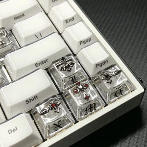 Könnte beinhalten: Nahaufnahme einer weißen mechanischen Tastatur mit individuellen Tastenkappen. Die Tastenkappen haben transparente Oberteile mit silbernen Kreuzen und roten Akzenten. Die Tastatur hat einen weißen Rahmen.