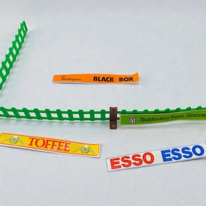 Puede incluir: Una valla de plástico verde para un juego de fútbol de mesa con postes de soporte marrones. Se incluyen tres pegatinas rectangulares: una naranja que dice Waddingtons BLACK BOX, una amarilla que dice TOFFEE y una blanca que dice ESSO ESSO.