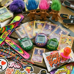 May include: A selection of 90s memorabilia, featuring Troll dolls, trading cards, Tamagotchi digital pets, stickers, mini skateboards, and a Ring Pop sweet. The items are displayed on a wooden surface.
