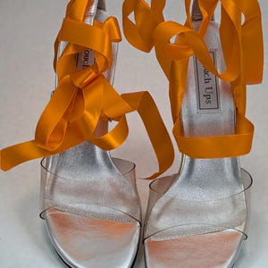 Könnte beinhalten: Ein Paar silberne High-Heel-Sandalen mit transparenten Riemen und leuchtend orangefarbenen Bändern. Die Sandalen haben eine metallisch silberne Innensohle und die Worte "Touch Ups" sind auf der Innensohle aufgedruckt.