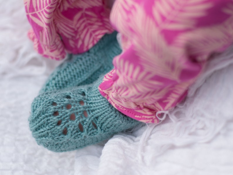 May include: A pair of teal blue knitted baby booties with a lacy pattern. The booties are on a white surface.