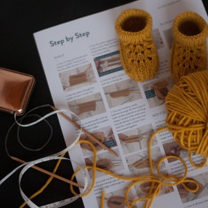May include: A close-up of a knitting project with instructions on a white sheet of paper. The project is a pair of yellow knitted baby booties. There is a ball of yellow yarn, two knitting needles, and a measuring tape.