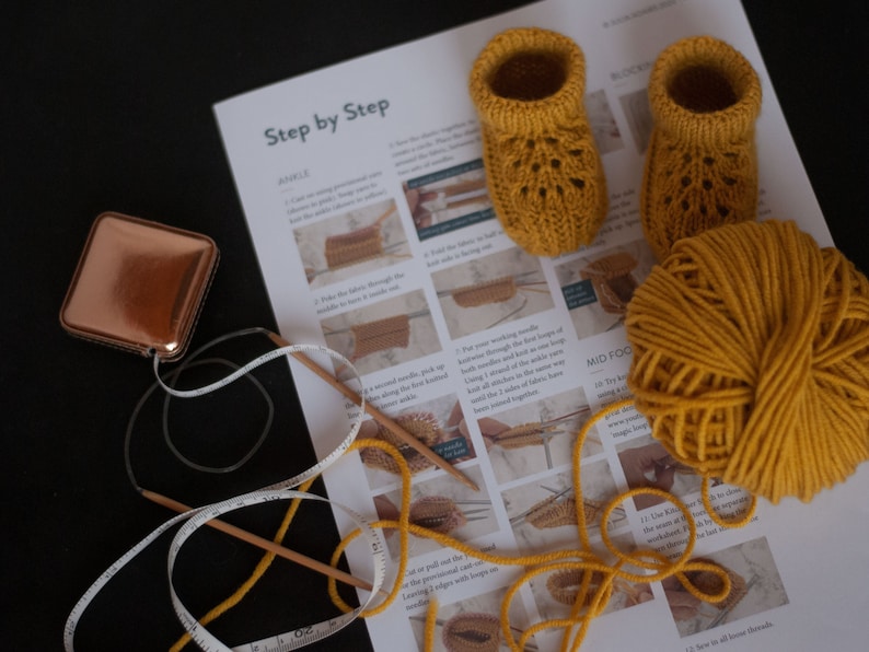 May include: A close-up of a knitting project with instructions on a white sheet of paper. The project is a pair of yellow knitted baby booties. There is a ball of yellow yarn, two knitting needles, and a measuring tape.