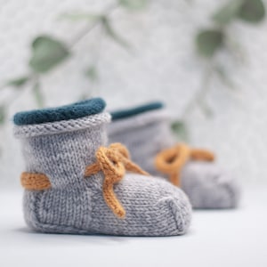 May include: A pair of gray knitted baby booties with a teal blue cuff and a yellow bow. The booties are on a white surface.