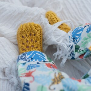 May include: A pair of yellow knitted baby booties with a lacy pattern. The booties are on a white blanket with a baby wearing a white and blue patterned onesie.