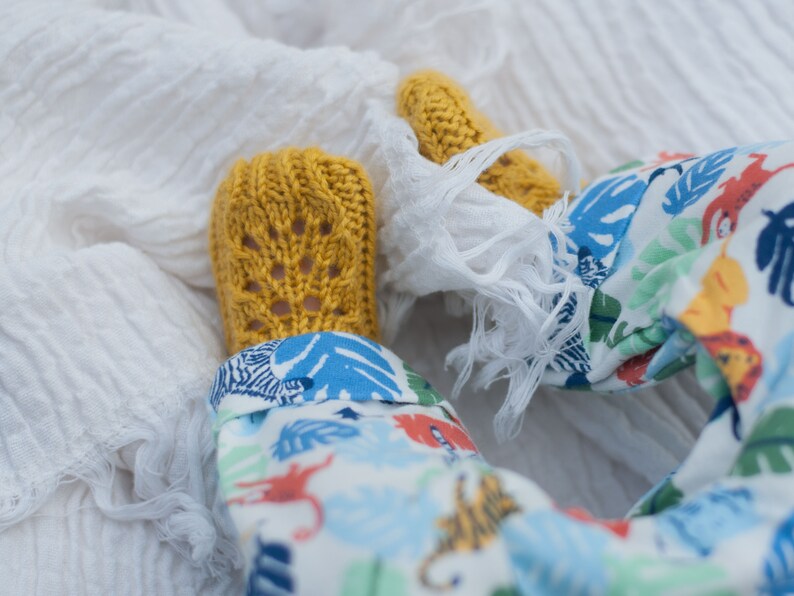 May include: A pair of yellow knitted baby booties with a lacy pattern. The booties are on a white blanket with a baby wearing a white and blue patterned onesie.